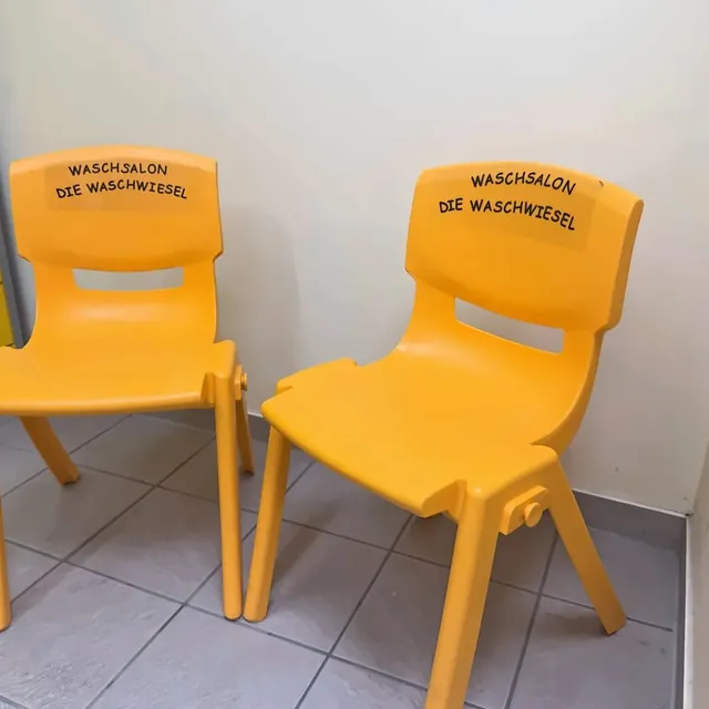 Zwei gelbe Stühle mit dem Schriftzug „Waschsalon Die WaschWiesel“ auf der Lehne