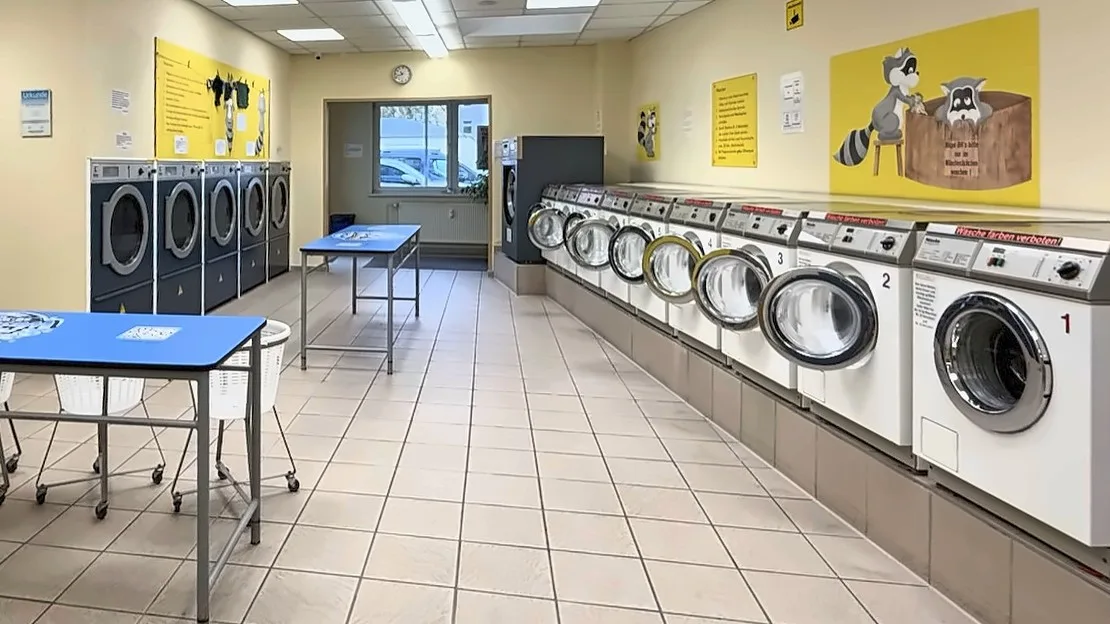 Blick in den Waschsalon: rechts eine Reihe weißer Miele-Waschmaschinen, links dunkle Trockner und blaue Falttische
